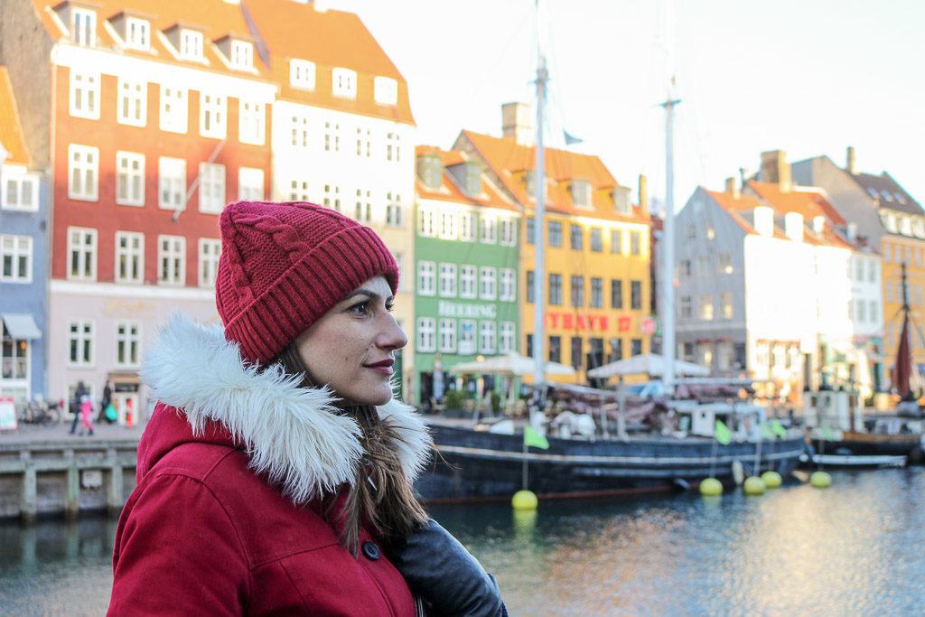 Photo of a woman wearing a red jacket and hat in front of the colored building in Copenhagen, Denmark's capital city. A great city to add to your European trip itinerary.