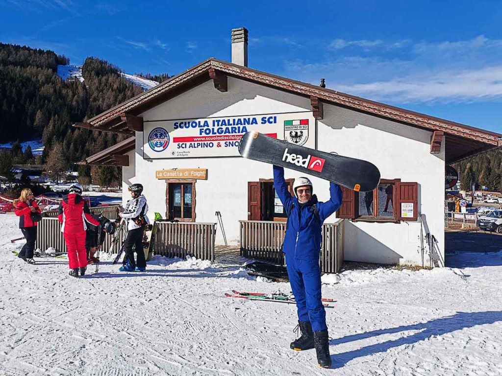 Man lifting a snowboard in front of winter sports school at Folgaria Lavarone ski area.