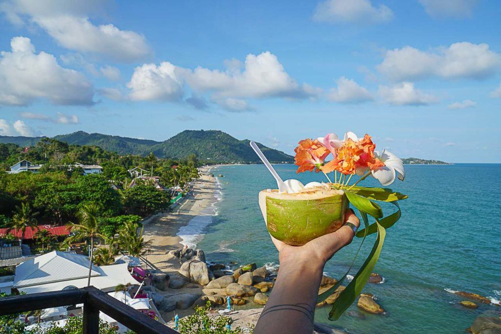 Mão segurando um drink de coco decorado com flores sobre uma praia na Tailândia. Descubra com o Love and Road as melhores maneiras de viajar para Koh Samui.