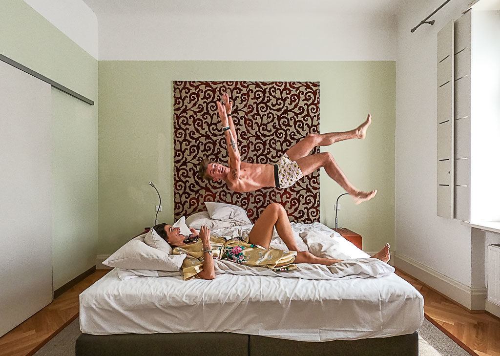 Man jumping on a hotel bed in Europe while the woman is lying on the bed and laughing.