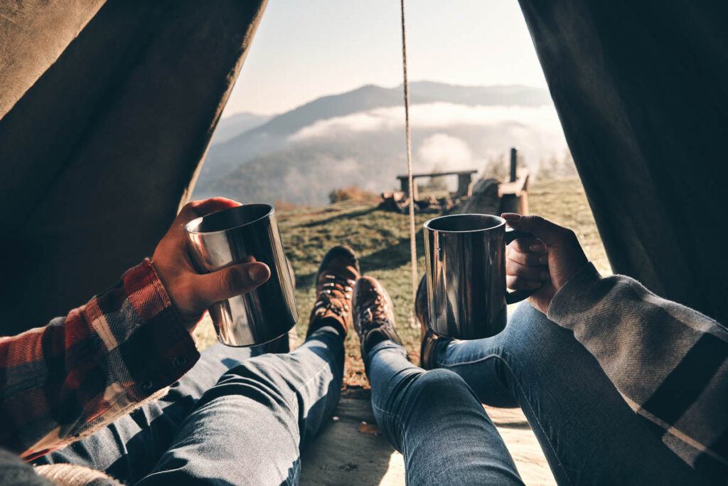 Fresh coffee to continue the adventure. Close-up of a young couple holding mugs while enjoying the view of a mountain range from their tent. Buying a good tent is the best thing you can do to do several road trips on a budget. 