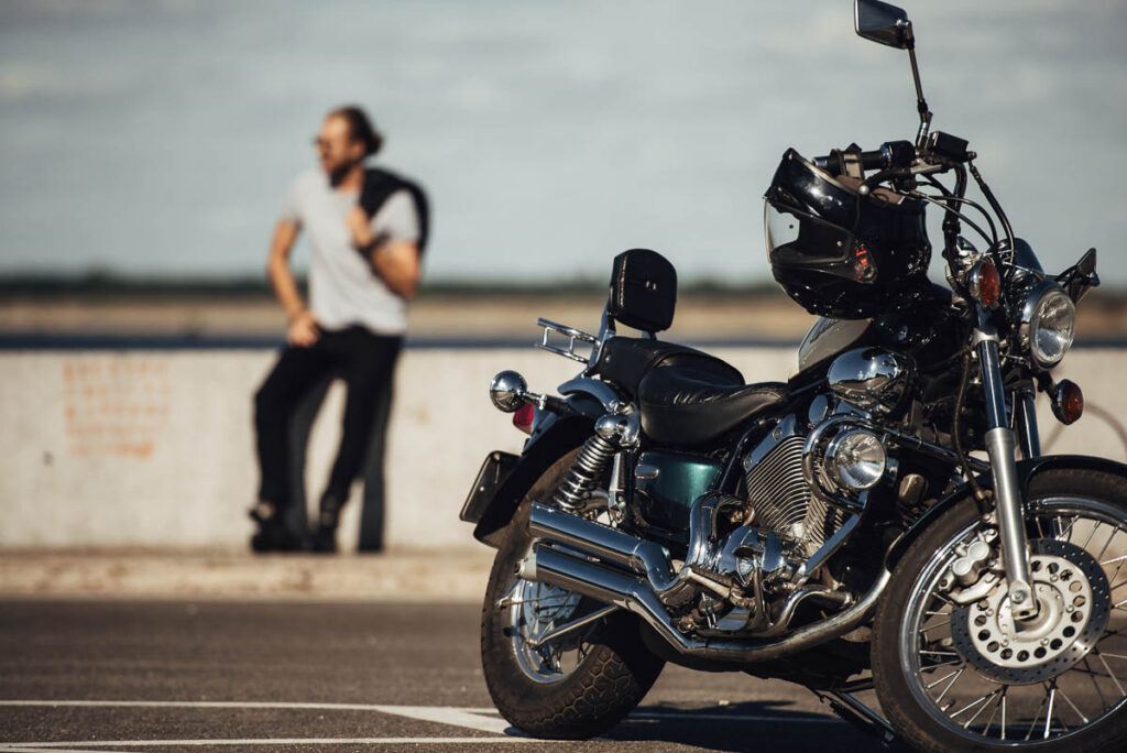 Photo of a motorbike parked on the side road and a motorcyclist in the background resting. 