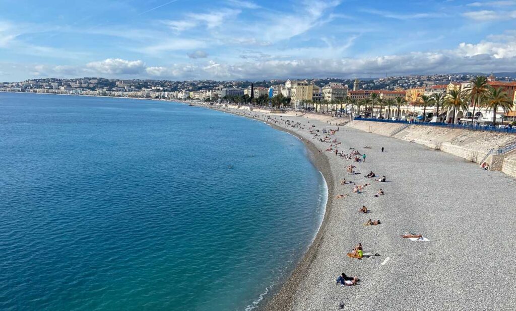 Beach in Nice, France. It's a pebble beach with crystal clear blue water. in the back is the city, a promenade with palm trees and historical buildings.