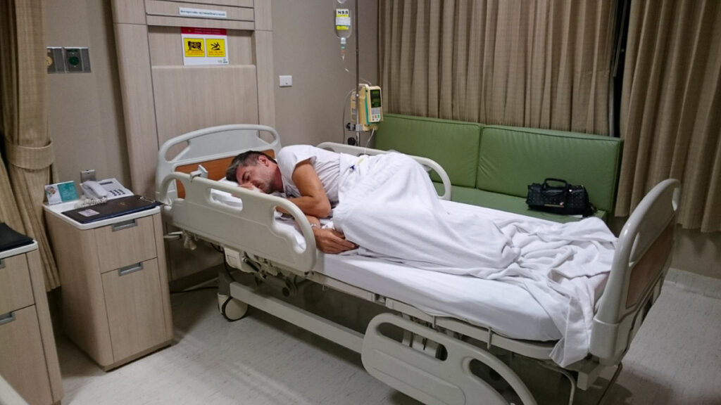 Rob deitado em uma cama de hospital em Bangkok. Ele ficou muito doente e precisou de cuidados médicos. É muito importante ter um seguro viagem para Bangkok, Tailândia.