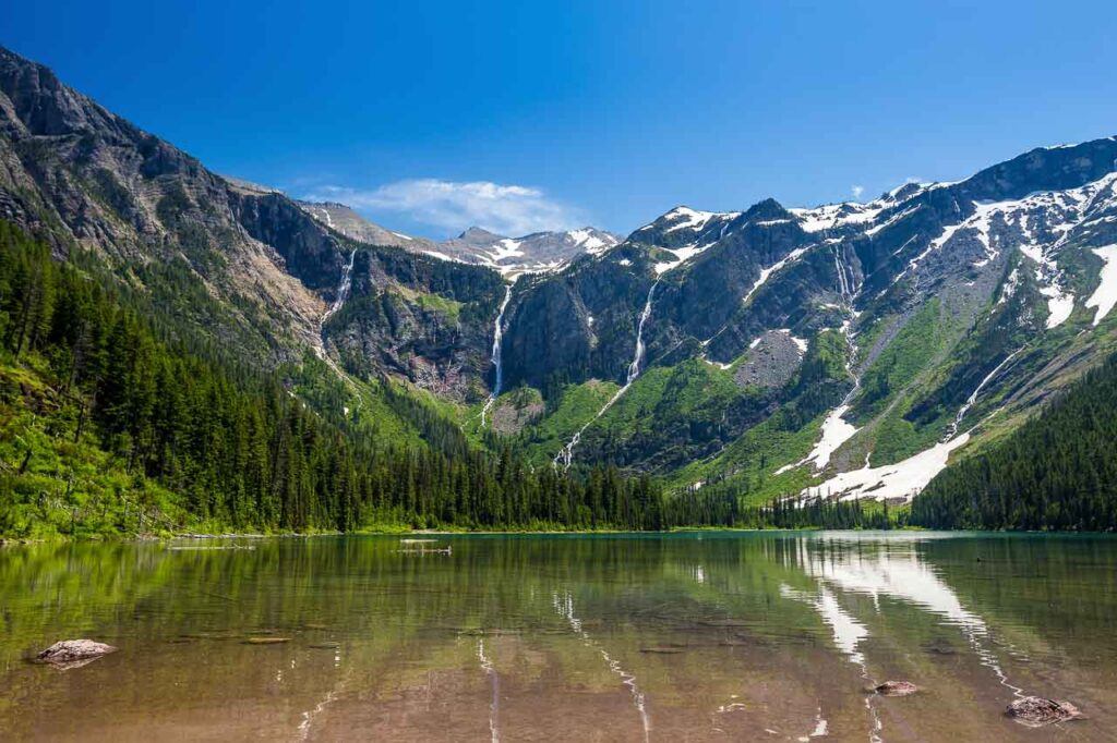Scenic views of Avalanche Lake in Glacier National Park, Montana, USA. The mountain range is in the back, with hills covered in the green forest and a lake in front.
