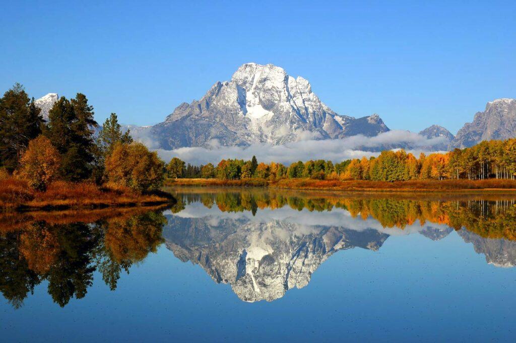 Reflection of the mountain range in a lake at Grand Teton National Park. The mount s covered in snow, and the forest around the lake has reddish autumn colors.