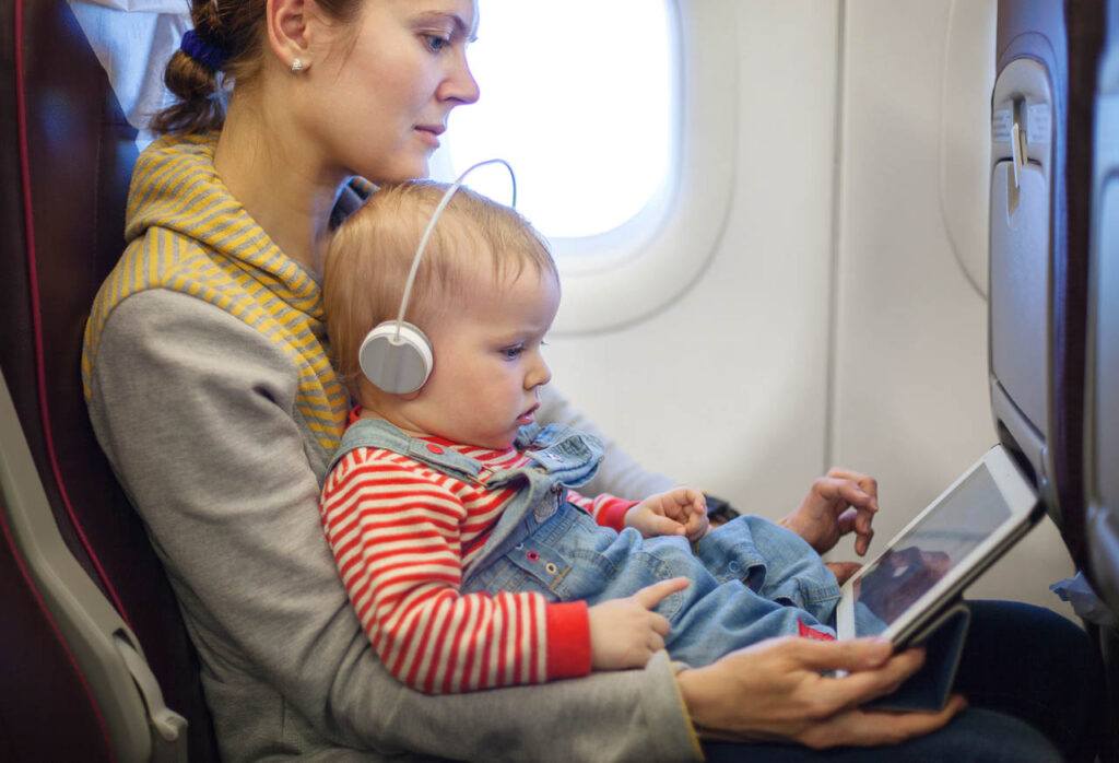 A mother with a small child sitting inside an airplane, she is entertaining the kid with a tablet.