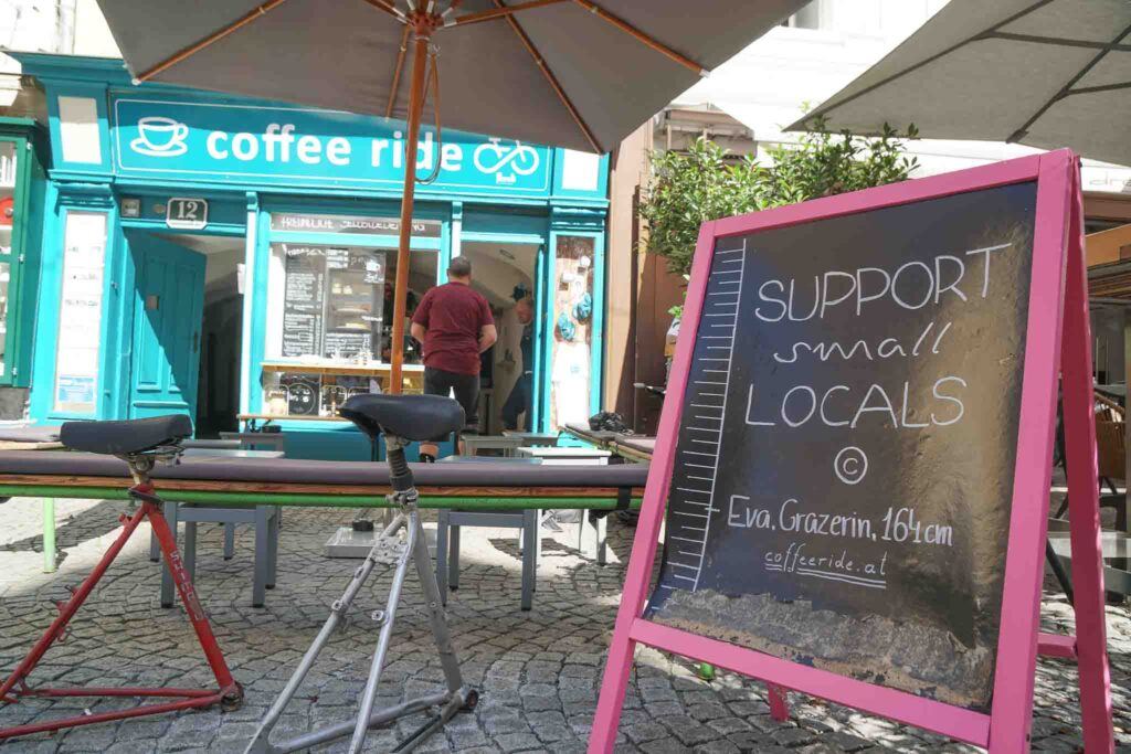 Placa na frente da cafeteria dizendo "apoie os pequenos comerciantes". Este é um exemplo de turismo sustentável em Graz na Áustria. 
