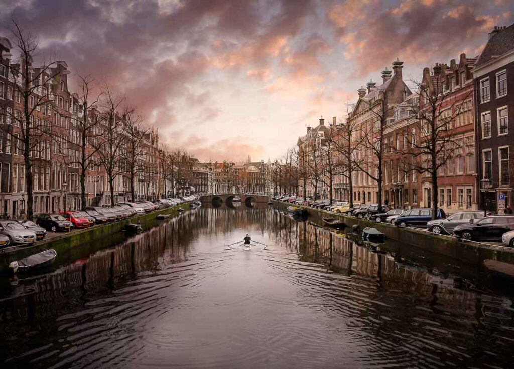 Photo of a canal in Amsterdam, Netherlands. It shows a kayak in the canal and during sunset, surrounded by beautiful canal houses. The photo was taken during Amsterdam's winter season. 