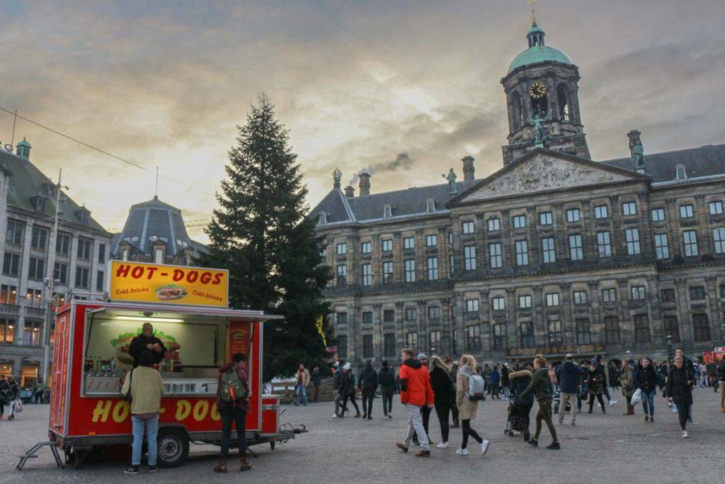 Photo of the Dam Square in Amsterdam. It shows the Royal Palace in the background, a hotdog food truck and a Christmas tree.