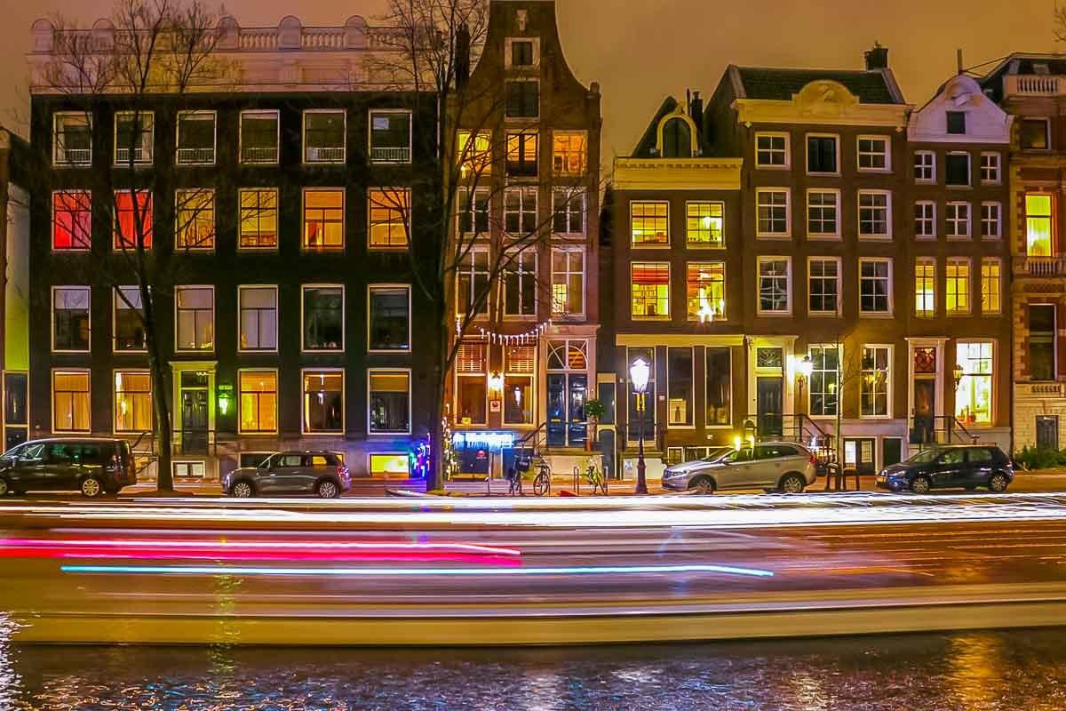 Photo of canal houses in Amsterdam in winter. It's nighttime and the widows are lit up with boat lights crossing in front of them.