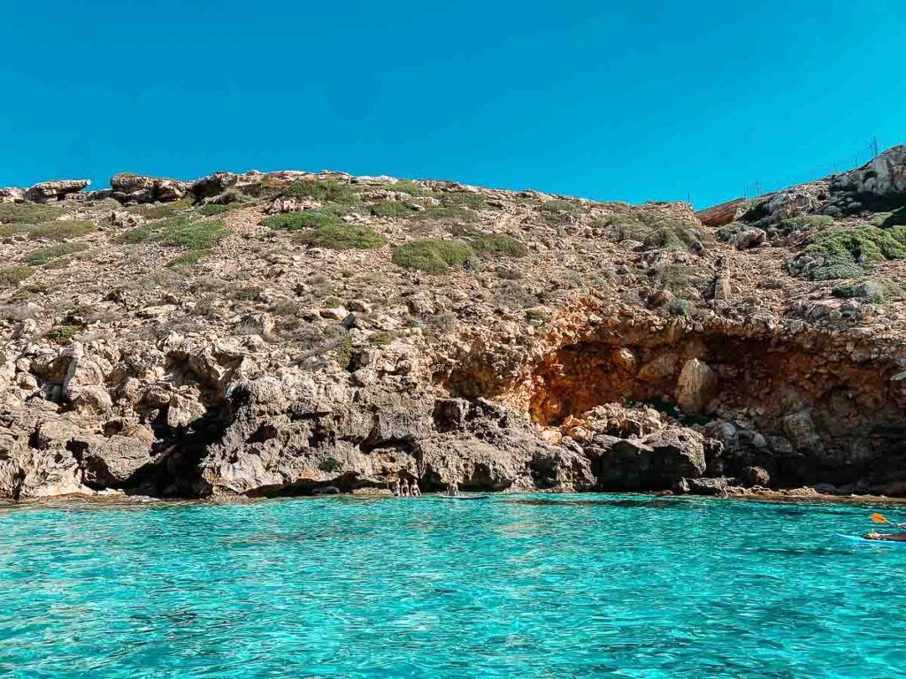 A beach cove on Mallorca Island, the turquoise water contrasts with the rock in the background. A few people are swimming in the cove.